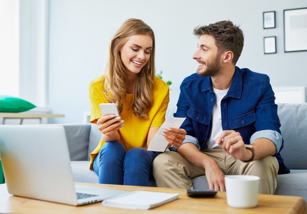 Happy couple looking at electricity bill and phone while sitting on a couch.